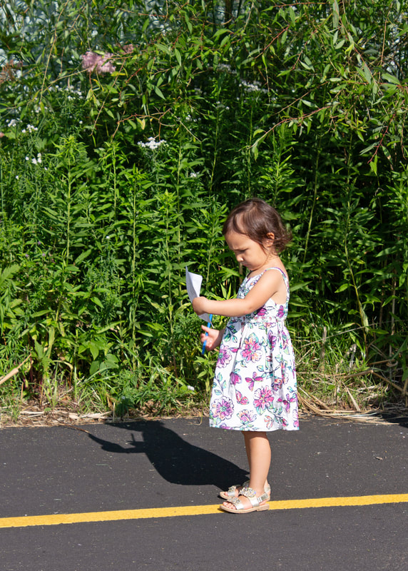 A young child standing outdoors next to various plants, holding a scavenger hunt checklist and concentrating very hard.