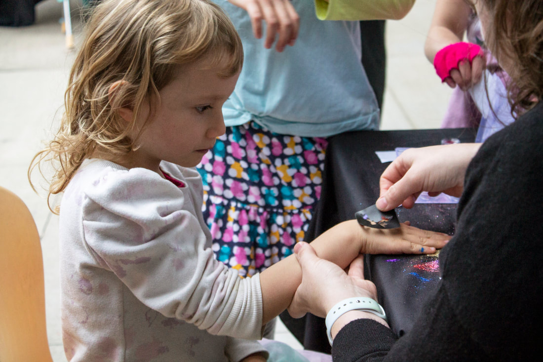 Outdoors, the glitter tattoos station is assisting a student with a hand tattoo by placing the sticker to their skin before beginning. The child is focusing hard on the volunteers work.