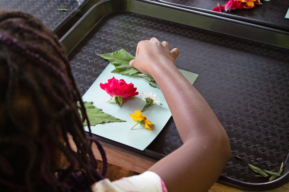 Looking over the shoulder as a student works on placing various flowers and leaves on a paper on a black tray.