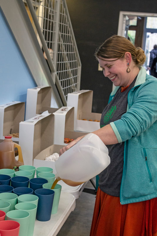 Lead guide assists with preparing glasses of apple cider for the event into blue, light blue, and red cups. She is standing in front of many boxes of donut holes.