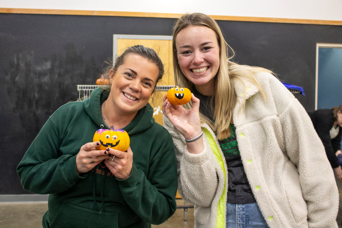 Two TMS staff members are seen holding up mini decorated pumpkins they created themselves. Behind them is a black board. The person to the left is wearing dark green and the person to the right is wearing white.