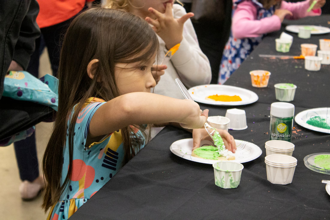 Child works to decorate a cookie with green frosting while focusing on a cookie across the table. Another student is show licking frosting from their finger as they finished eating their cookie.