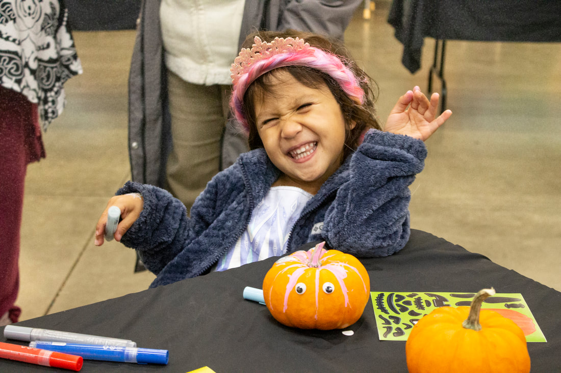 Smiling with much excitement, a student is happily leaning back wearing a fuzzy coat and pink crown. They are holding a paint marker with a half decorated mini pumpkin sitting in front of them on a black table with other paint markers.
