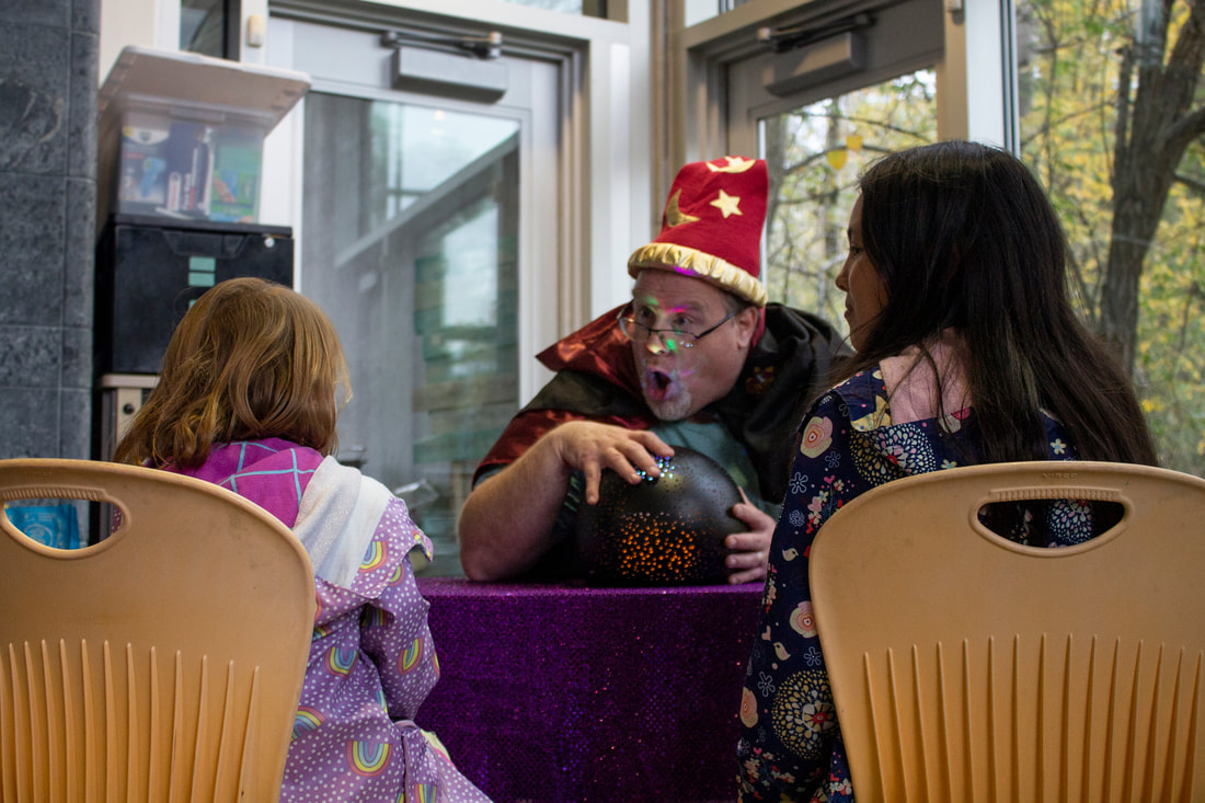 A volunteer dressed as a psychic is shown dramatically telling a fortune to a student while their hand waves over their crystal ball. Different colors of light are reflecting off of the volunteer's face from the ball. The student getting the reading is facing the volunteer wearing a light purple raincoat with rainbows and the other student is watching the student to the left's reaction to the news. They are wearing a dark green coat with many different types of flowers.