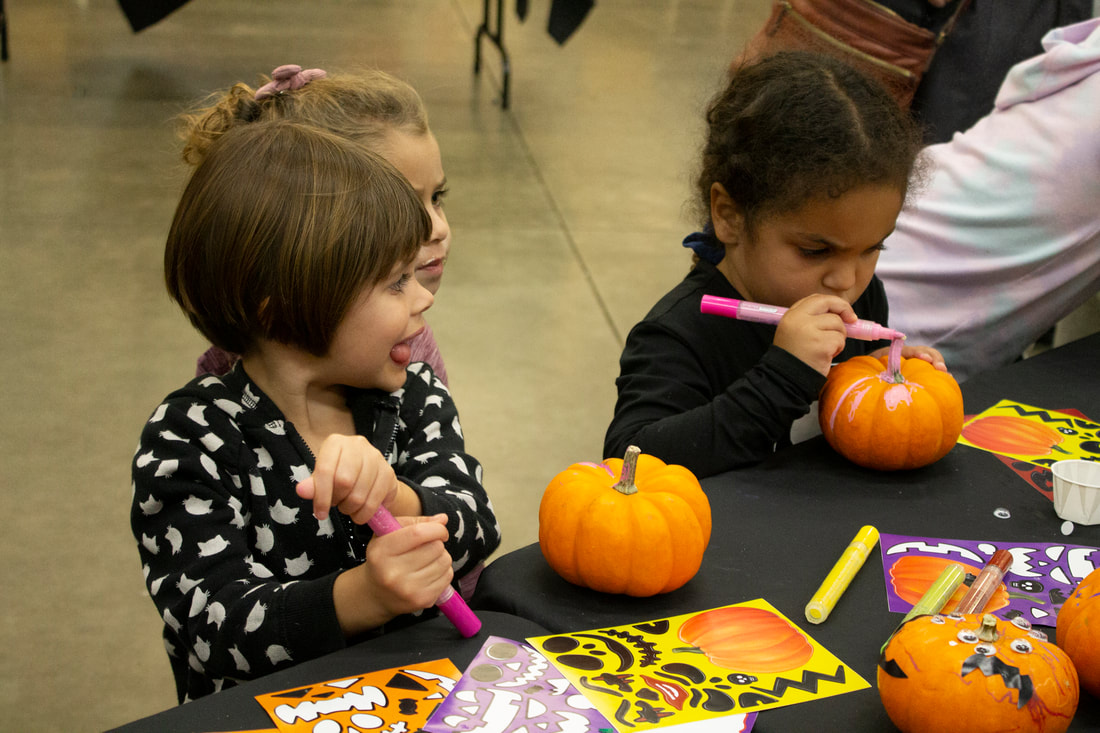 Three students gather at the mini pumpkin decorating table. One is hard at work coloring the pumpkin, another has stepped back to look around the room, and the third has their tongue out sideways as they hold a paint marker with both hands and ponder how they will start on their pumpkin.